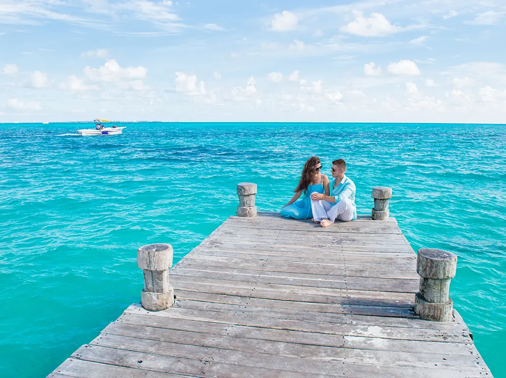 Young beautiful couple in blue and white clothes are sitting at the wooden pier. Turquoise color water behind them. Vacation